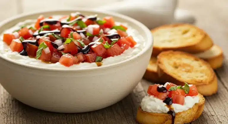 Overhead view of a party platter with a bowl of bruschetta dip surrounded by crackers, carrots, and cucumber slices.