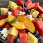 A close-up photo of a glass bowl of summer peach fruit salad, glistening with honey-lime dressing. The colorful medley includes sliced peaches, strawberries, blueberries, watermelon, pineapple, crumbled feta cheese, and fresh mint.