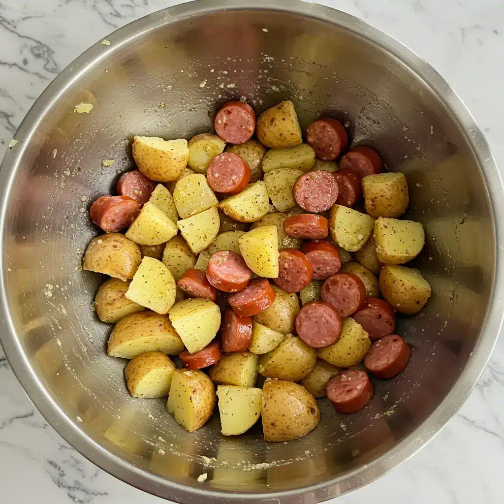 An overhead shot of a stainless steel bowl with cubed potatoes, sliced sausage, and ranch seasoning mixed together before baking.