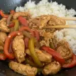 A close-up of a white bowl filled with homemade chicken and pepper stir fry served over fluffy rice, with chopsticks lifting a piece of chicken.