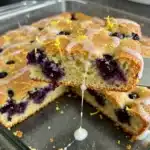 A stunning, moist Lemon Glazed Blueberry Boyfriend Bait cake, drizzled with bright lemon glaze, on a white cake stand, with fresh blueberries scattered around. A perfect homemade dessert by Karen Colman.