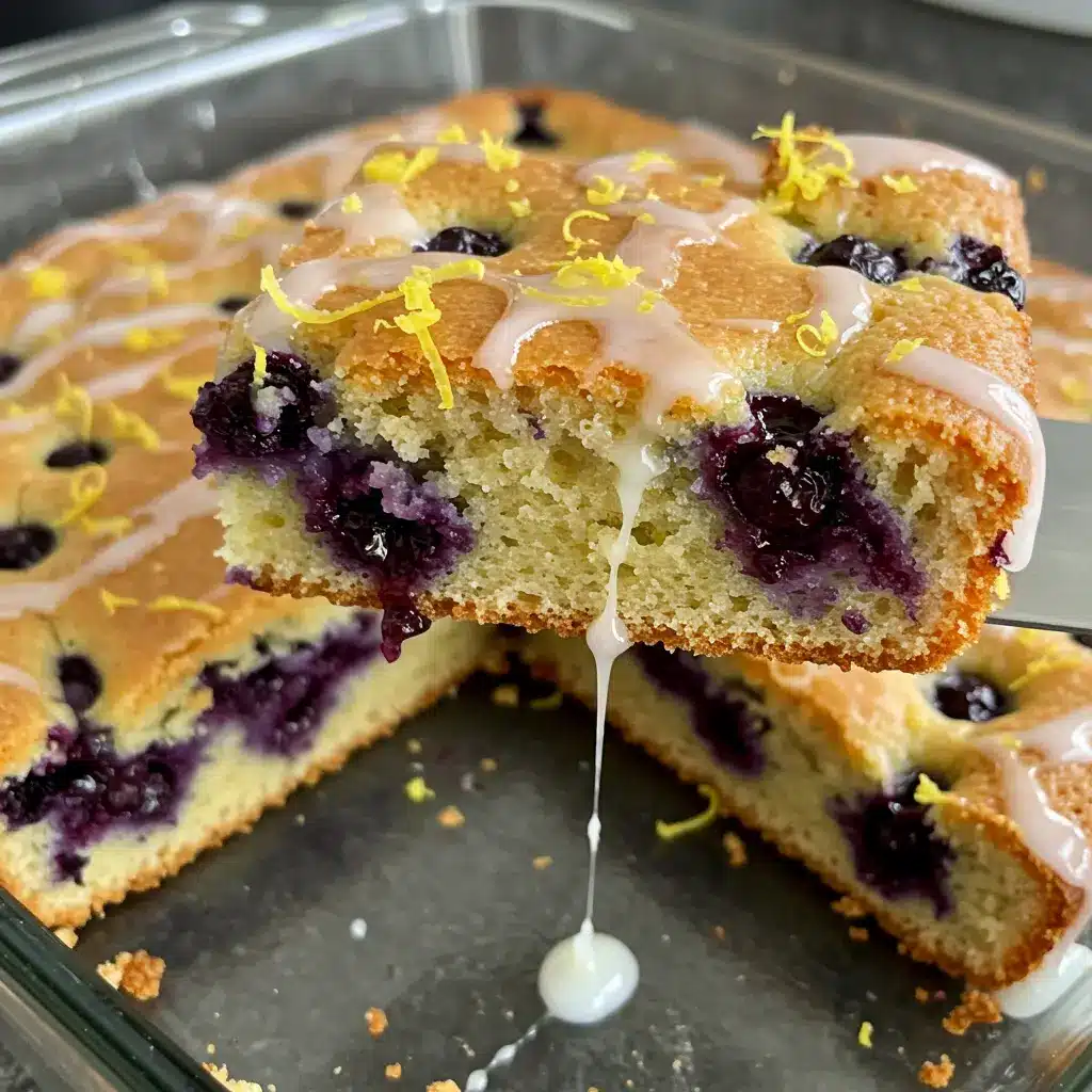 A stunning, moist Lemon Glazed Blueberry Boyfriend Bait cake, drizzled with bright lemon glaze, on a white cake stand, with fresh blueberries scattered around. A perfect homemade dessert by Karen Colman.