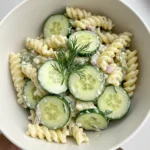 A close-up overhead shot of a white bowl filled with creamy fresh cucumber pasta salad, with thinly sliced cucumbers and fresh dill arranged on top.