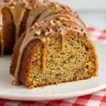 A moist slice of pecan zucchini bread on a white plate, with a thick caramel-pecan glaze dripping down the side and visible flecks of zucchini in the crumb.
